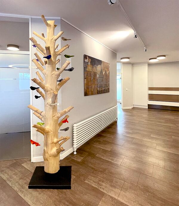 Blick Praxiseingang - vorne steht ein künstlich erstellter Baum aus Holz, daran hängen bunte Vögel, gebastelt aus Filz